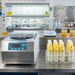 A professional food science laboratory bench photographed in bright, clinical lighting. A refrigerated benchtop centrifuge is running in the foreground, its digital display showing temperature and speed settings. 