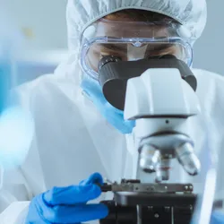 Researcher focused on microscope, wearing protective equipment.