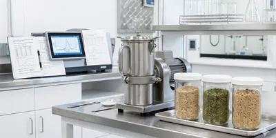  food science laboratory workbench showing a stainless steel knife mill next to several labeled sample containers filled with homogenized food matrices — ground grain, dried herbs, and a composite meal sample.