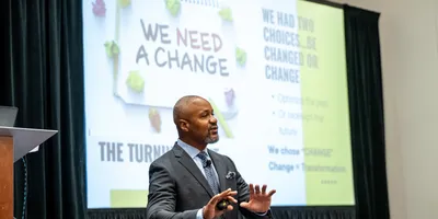 Curtiss McNair in suit standing at the front of a conference room delivering his 2026 Lab Manager Leadership Summit keynote, with slide deck in the background