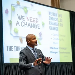 Curtiss McNair in suit standing at the front of a conference room delivering his 2026 Lab Manager Leadership Summit keynote, with slide deck in the background