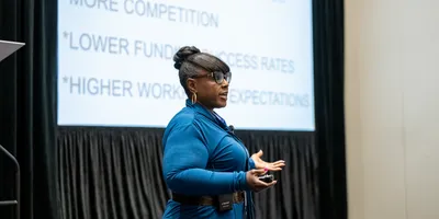 Tarshae Drummond wearing a blue jumpsuit, standing in front of a large screen displaying a slide for her presentation, and delivering the presentation to a room of attendees.