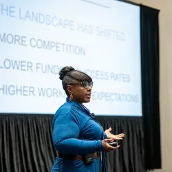 Tarshae Drummond wearing a blue jumpsuit, standing in front of a large screen displaying a slide for her presentation, and delivering the presentation to a room of attendees.