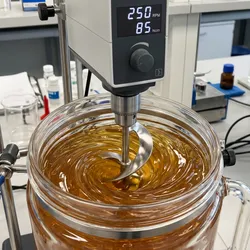 An overhead lab mixer using a helical ribbon impeller to achieve blend uniformity in a high-viscosity amber gel sample, illustrating efficient laminar flow.