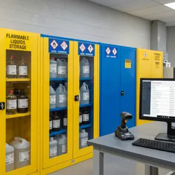 A photorealistic image of a laboratory chemical storage area with clearly labeled containers of various hazard classes arranged in segregated storage cabinets, a barcode scanner on a bench surface nearby, and a computer monitor displaying a chemical inventory management interface.