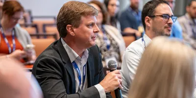 A male attendee in a suit, holding a microphone to pose a question about leading with influence