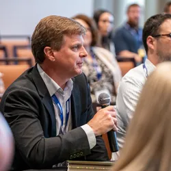 A male attendee in a suit, holding a microphone to pose a question about leading with influence