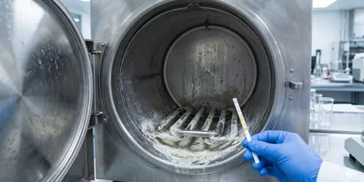 A technician’s gloved hand holds a water quality test strip in front of an open stainless steel autoclave chamber showing significant mineral scale buildup on its heating elements.