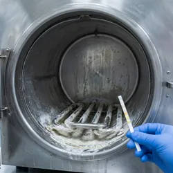 A technician’s gloved hand holds a water quality test strip in front of an open stainless steel autoclave chamber showing significant mineral scale buildup on its heating elements.