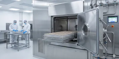 An open pharmaceutical-grade stainless steel autoclave chamber reveals a large rack loaded with rows of glass vials, while two fully gowned technicians work at a table in the background of a bright cleanroom environment.
