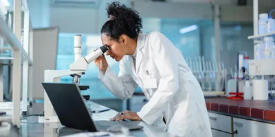 Young Hispanic scientist wearing a lab coat, looking under microscope while using laptop in a laboratory.