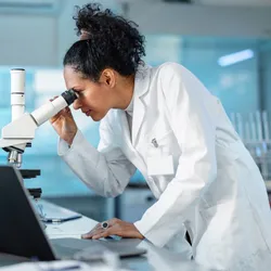 Young Hispanic scientist wearing a lab coat, looking under microscope while using laptop in a laboratory.