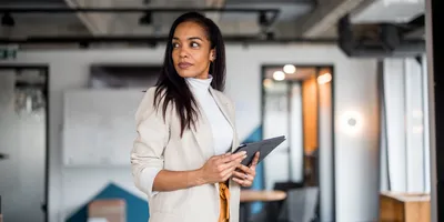 A mature Hispanic woman in professional business attire holds a digital tablet in a modern office setting.