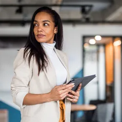 A mature Hispanic woman in professional business attire holds a digital tablet in a modern office setting.
