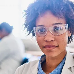 Portrait of a confident young female scientist sitting a table in a lab with colleagues working in the background