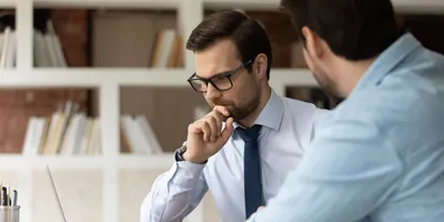 Thoughtful young male boss leader in eyewear looking at laptop screen, discussing project problem solution with colleague at brainstorming meeting
