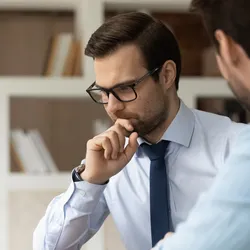 Thoughtful young male boss leader in eyewear looking at laptop screen, discussing project problem solution with colleague at brainstorming meeting