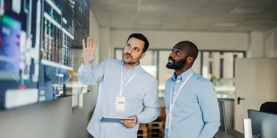 Two adult male engineers, a mid adult and a young adult, collaborating and discussing crucial data visualization on a large digital screen in a modern control room