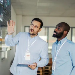 Two adult male engineers, a mid adult and a young adult, collaborating and discussing crucial data visualization on a large digital screen in a modern control room