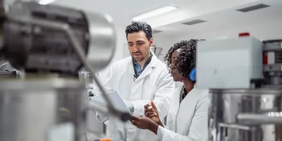 Close-up portrait of young multi-ethnic pharmaceutical professionals in white lab coats examining documentation near industrial laboratory equipment.