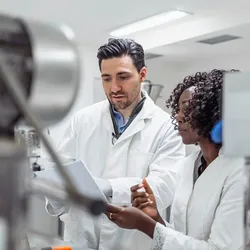 Close-up portrait of young multi-ethnic pharmaceutical professionals in white lab coats examining documentation near industrial laboratory equipment.