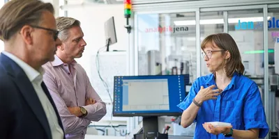 Luzia Gyr holds a microplate while explaining JenXplor laboratory automation processes to Lars Böttcher and Sascha van Beek in front of a robotic workstation.