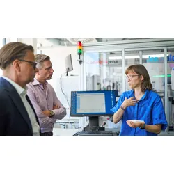 Luzia Gyr holds a microplate while explaining JenXplor laboratory automation processes to Lars Böttcher and Sascha van Beek in front of a robotic workstation.