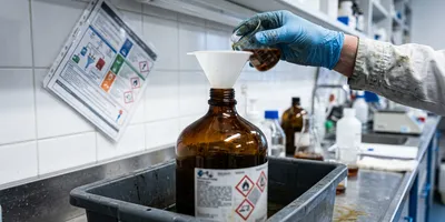 A gloved hand pouring from a beaker into a funnel seated in an amber solvent waste carboy standing in a secondary containment tray
