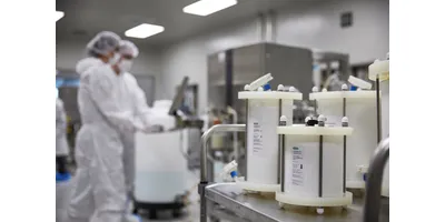 Foreground view of white prepacked chromatography columns on a stainless steel cart, with two fully gowned technicians operating equipment inside a sterile GMP cleanroom behind them.