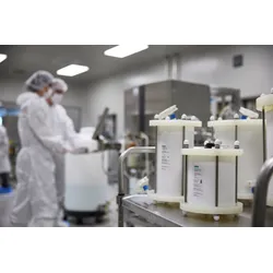 Foreground view of white prepacked chromatography columns on a stainless steel cart, with two fully gowned technicians operating equipment inside a sterile GMP cleanroom behind them.