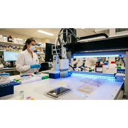 Robotic multi-channel liquid handler dispensing into 96-well plates for enzymatic DNA synthesis, with a scientist monitoring at a computer in the background.