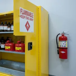 An open laboratory flammables storage cabinet holding amber solvent bottles and red safety cans, with a wall-mounted fire extinguisher alongside