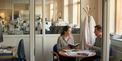 Two colleagues seated at a round table in a glass-walled laboratory office, one listening with a notebook while the other speaks