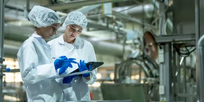 Two scientists wearing protective gear and gloves using a digital tablet in an industrial laboratory environment.