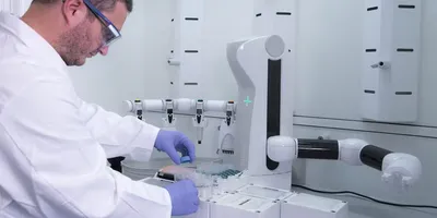 A scientist in a white lab coat and blue nitrile gloves loads labware onto an Andrew+ Robot inside an Andrew+ Pod enclosure in a pharma QC lab.
